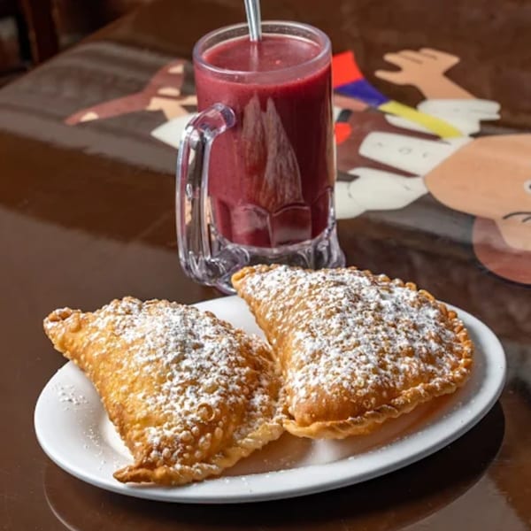Two golden-fried empanadas dusted with powdered sugar, next to a glass of red juice.