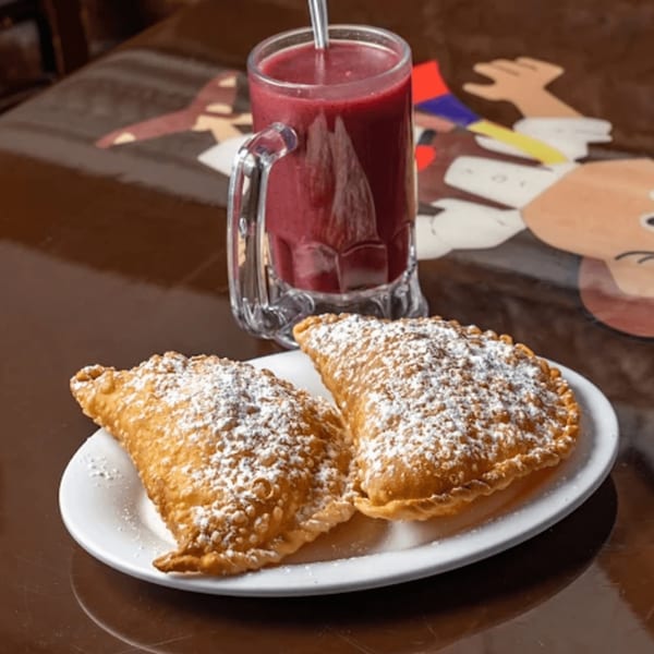 Two powdered sugar-dusted empanadas on a white plate with a red drink in a mug.