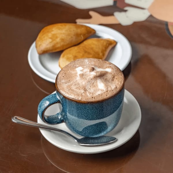 A blue mug of hot chocolate with foam art and two golden-brown Colombian empanadas on a white plate.