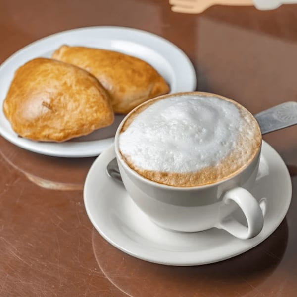 A cup of frothy cappuccino and two golden-brown Colombian empanadas on a white plate.