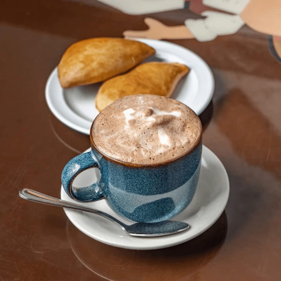 A blue mug of hot chocolate with foam art and two Colombian empanadas on a white plate.